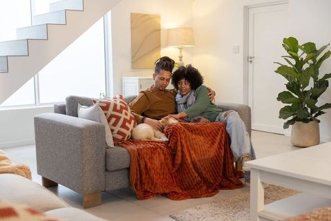 African american couple cuddling on sofa with dog under rust blanket in cozy living room