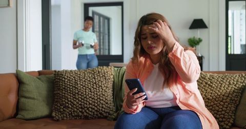 Stressed african american woman sitting on sofa checking phone while friend bringing coffee