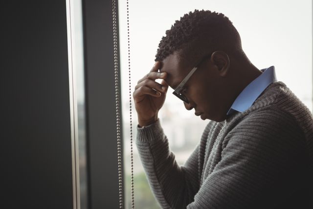 Businessman standing by office window, appearing stressed and deep in thought. Useful for illustrating workplace stress, mental health in corporate settings, and professional challenges.