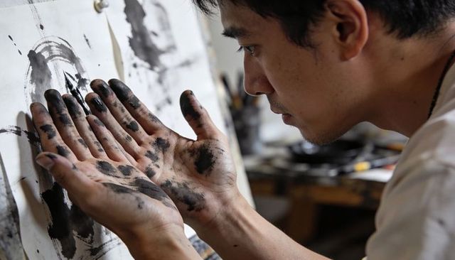 Asian artist examining ink-stained hands against canvas, intimate studio process