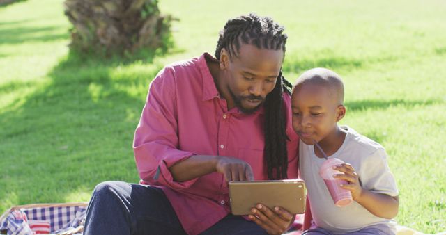 Father and son spending quality time together in a park, using a tablet device while the son enjoys a drink. Perfect for content related to family bonding, outdoor activities, and technology usage among children.