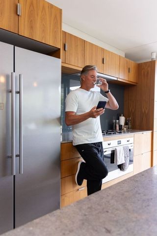 Mature man leaning in modern kitchen checking smartphone while drinking glass of water