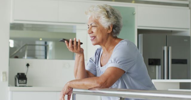 This image depicts an elderly woman using voice command on her smartphone while leaning on a railing in a modern kitchen. She appears relaxed and cheerful, suggesting a comfortable and familiar use of technology in her daily life. This visual can be used for advertising tech products targeting seniors, illustrating ease of use, promoting independence in elderly, or highlighting modern living conditions for older adults.