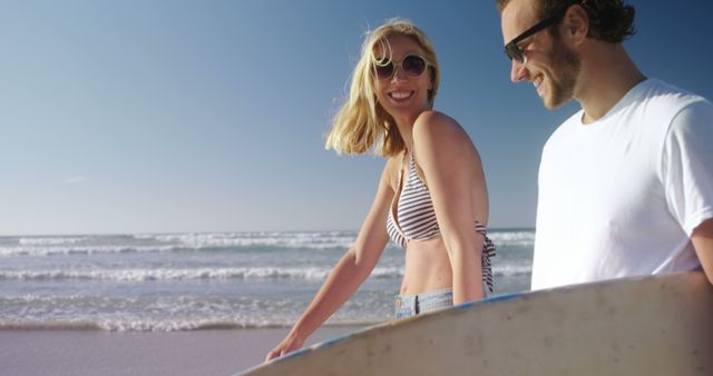 A young Caucasian woman and man carry a surfboard on a sunny beach, with copy space. Their casual attire and smiles suggest a leisurely day of surfing and relaxation by the sea.