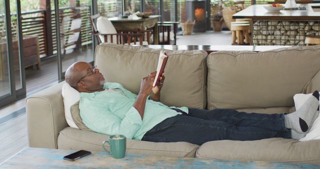 Man lying comfortably on a couch reading a book. Cozy indoor environment suggesting leisure time or relaxation at home. Could be used for themes like reading, relaxation, indoor activities, home lifestyle, or comfort.