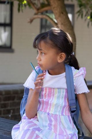 Thoughtful schoolgirl sitting on bench with pastel dress and backpack, holding pencil