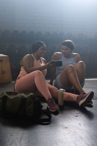 Two african american women resting in gym studio activewear, sharing phone after workout