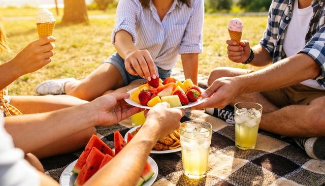 Friends sharing fresh fruit and lemonade on checked picnic blanket during golden hour