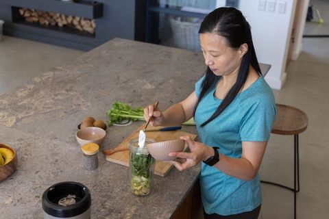 Mature asian woman preparing green smoothie with avocado and chia at kitchen island