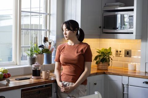 Asian woman gazing out kitchen window with french press on counter, tranquil morning light