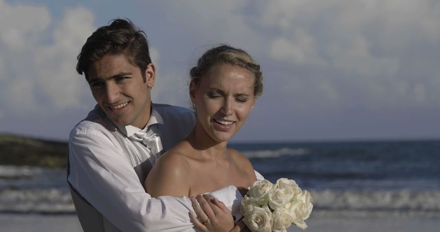 Happy newlywed couple embracing on beach with bouquet - Download Free Stock Images Pikwizard.com