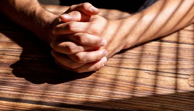 Clasping male-presenting hands resting on sunlit wooden table casting blinds shadows
