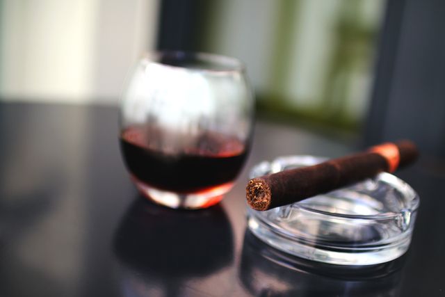 Close shot of a glass of whiskey next to a lit cigar resting in an ashtray on a polished black table. Image conveys themes of luxury, relaxation, and sophistication, making it ideal for use in marketing materials for upscale bars, cigar lounges, or lifestyle magazines.