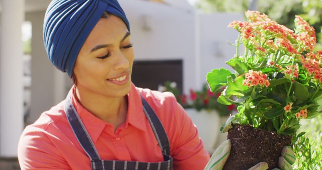 A woman with a blue turban and apron is planting flowers in a garden. She smiles while handling a potted plant. This image is ideal for use in articles or guides about home gardening, botanical hobbies, sustainability, or for promoting gardening supplies and tools.
