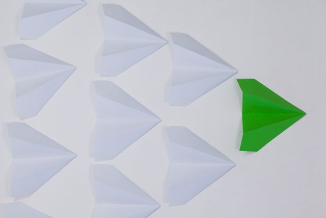 Green paper airplane leading a group of white paper airplanes on a white background. Ideal for concepts of leadership, innovation, and standing out. Useful for business presentations, motivational posters, and educational materials.
