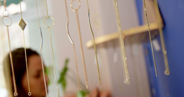 A young woman is seen in the background through a display of delicate hanging necklaces, with copy space. Jewelry is the focal point, suggesting a boutique or craft market setting where unique pieces are showcased.