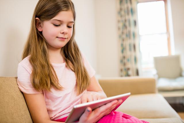 Young girl sitting on sofa using tablet in living room at home. Ideal for content related to children and technology, digital learning, home activities, and family life.