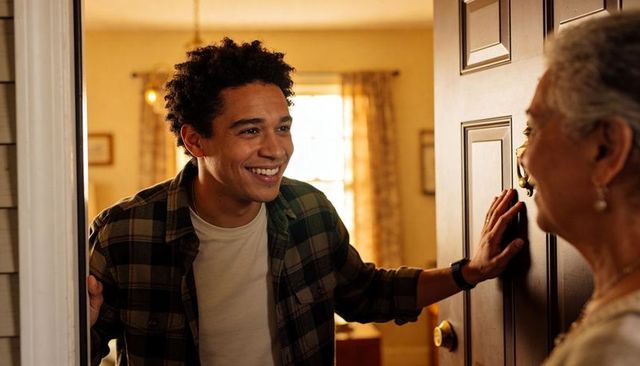 Smiling young man greeting older woman at open doorway, leaning in with warm welcome