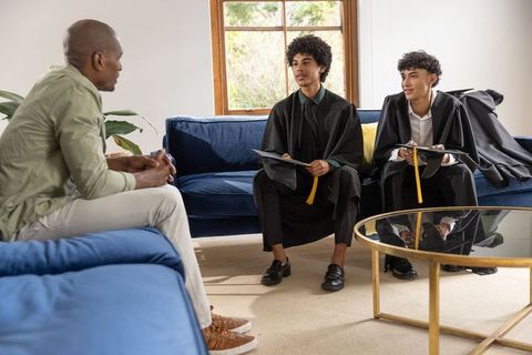 Two young graduates in caps and gowns holding yellow tassels talking to relative on sofa