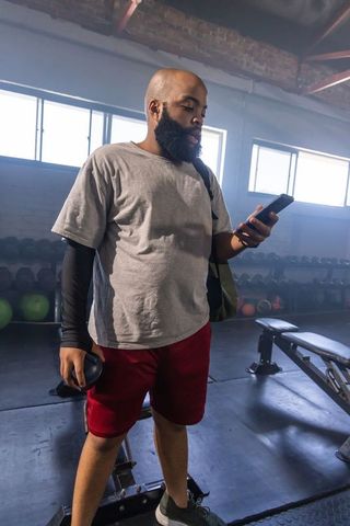 African american man checking smartphone after workout holding weight plate in urban gym
