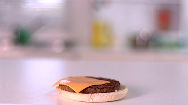 Slice of cheddar cheese is slowly landing on a beef burger patty atop a open-faced bun in a modern kitchen. This scene captures the moment just before assembling the burger, emphasizing freshness and quality ingredients. Perfect for culinary blogs, cooking tutorials, advertisements for gourmet burger restaurants, or cheese-related food campaigns.