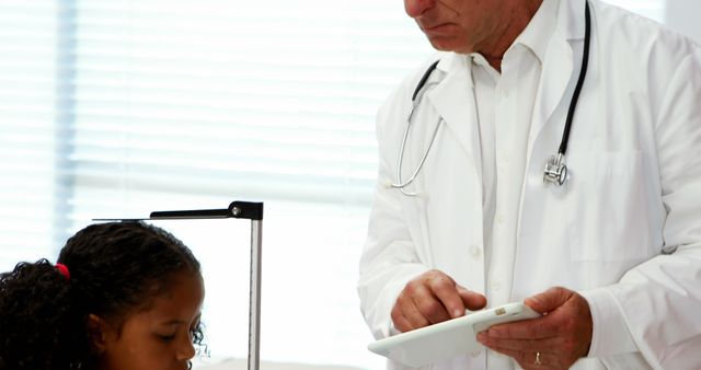 Male doctor, wearing white lab coat with stethoscope, using a tablet in modern clinic while examining young girl. Use this image for healthcare services, pediatric care promotions, medical technology integration, or clinic advertising.