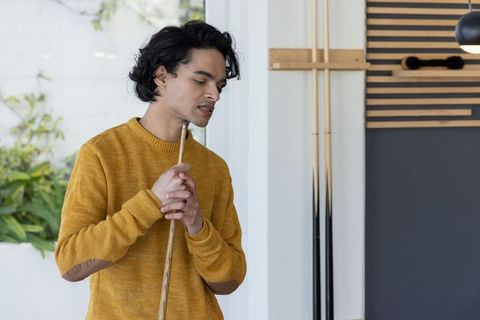 Contemplative asian man holding pool cue, chin near tip, wearing mustard sweater in game room
