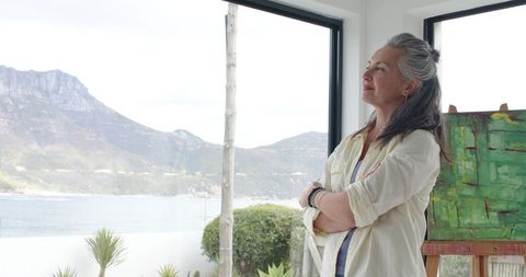 Mature woman standing with arms crossed contemplating coastal view in sunlit art studio