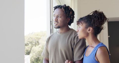 African american couple holding hands looking out sunlit window in modern apartment