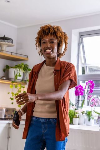 African american woman listening to music in kitchen wearing headphones holding smartphone