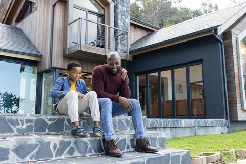 African american father and son sitting on stone steps of modern house using smartphone