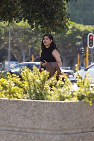 Woman standing by urban planter holding smartphone and tan coat on sunny sidewalk