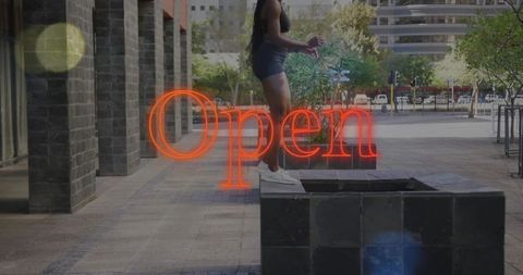 Urban athletic woman balancing on planter with neon Open sign in modern city plaza