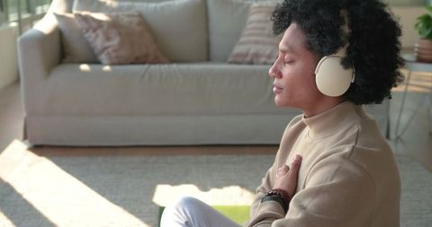 Mid-adult african american man meditating with over-ear headphones on mat in sunlit living room