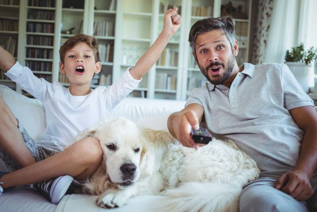 Father and son enjoying television together with their pet dog in a cozy living room. Ideal for use in advertisements or articles about family bonding, home life, parenting, leisure activities, and pet companionship.