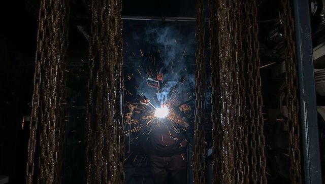 Welder welding amid hanging chains, creating bright sparks in dark industrial workshop