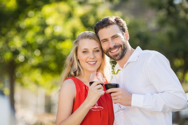 Happy couple holding wine glasses and toasting in a park. Ideal for use in advertisements, social media posts, or articles about relationships, outdoor activities, and celebrations.