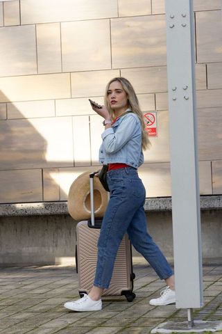 Young woman walking with rolling suitcase and straw hat speaking into phone in sunlit plaza