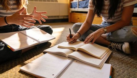 College students collaborating on dorm floor studying together with notebooks and textbook