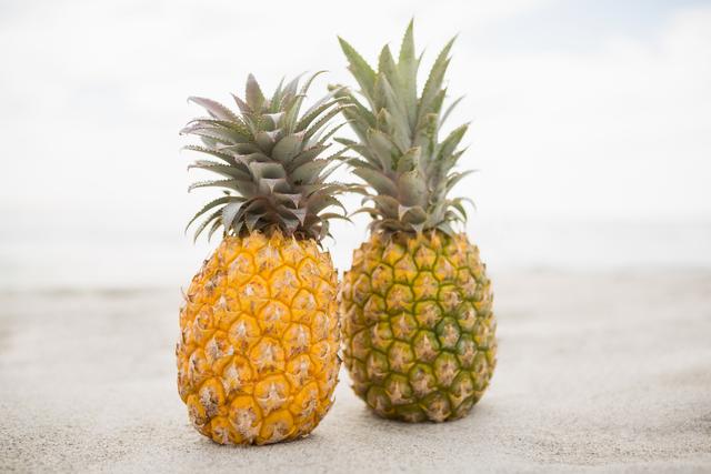 Two ripe pineapples are placed on sandy beach, evoking a tropical and exotic vibe. Ideal for use in travel brochures, summer vacation promotions, healthy eating campaigns, and tropical-themed designs. The image conveys freshness, relaxation, and the beauty of nature.