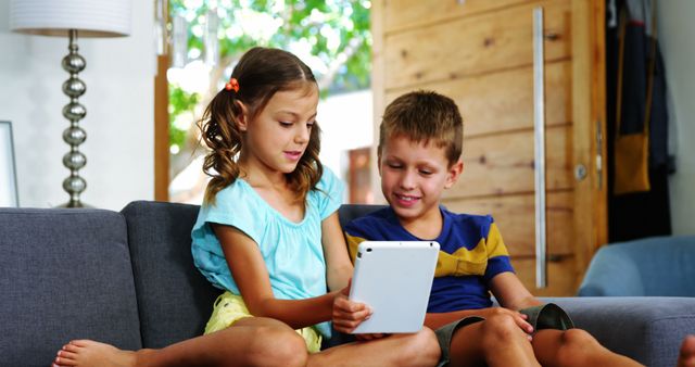 Young siblings sitting on sofa sharing and using a tablet together at home. A girl and a boy, wearing casual clothes, are engaged and focused on the screen, enjoying each other's company. This depicts a cozy and relaxed family environment. Suitable for illustrating family life, digital learning, or promotional content for technology products aimed at children.