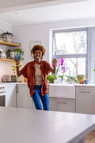 Joyful african american woman dancing in sunlit modern kitchen holding smartphone