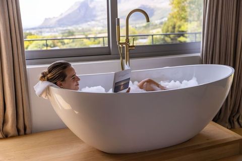 Woman relaxing in freestanding bathtub with bubble bath and mountain view reading booklet