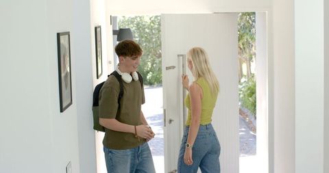 Teen boy returning home with backpack and headphones greeting woman at sunlit entryway