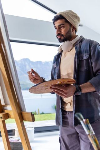 Indian artist painting lakeside mountain landscape at bright studio easel with headphones