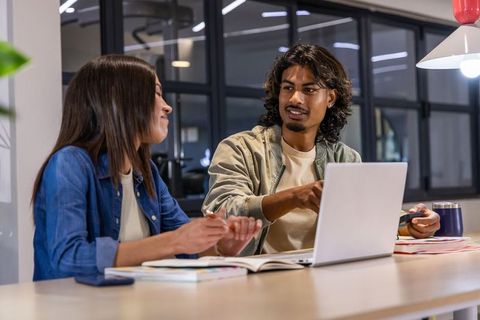 Indian man and Asian woman collaborating and pointing at laptop in modern coworking space