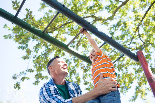 This image captures a joyful moment between a father and his son at a playground. The father is supporting his son as he plays on a jungle gym, highlighting themes of family bonding, support, and outdoor fun. Ideal for use in parenting blogs, family-oriented advertisements, or articles about childhood activities and outdoor play.