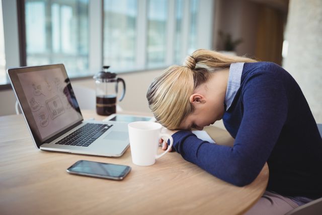 Businesswoman experiencing fatigue while working at desk in modern office. She is leaning her head on her arms next to a laptop, coffee cup, and smartphone. Ideal for illustrating workplace stress, professional burnout, or the need for work-life balance in corporate environments.