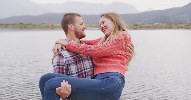 Picture shows happy couple embracing by a tranquil lake. Man is holding woman in his arms while they both smile lovingly. Scenic background with mountains and calm water. Suitable for themes of love, romance, outdoor activities, and serene natural settings.