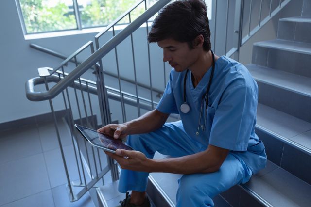 Caucasian male surgeon in blue scrubs sitting on hospital stairs using a digital tablet. Ideal for illustrating modern healthcare, medical technology, and the daily life of medical professionals. Suitable for use in healthcare blogs, medical websites, and educational materials.